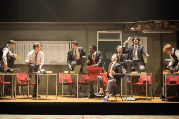 The boys from The History Boys dance in front of a classroom set.